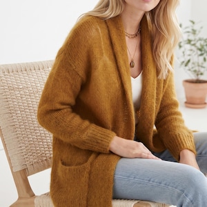 Peut inclure: Un cardigan ouvert jaune moutarde avec un col châle, des manches longues et deux poches avant. Le vêtement en tricot présente une surface texturée et duveteuse, porté sur un haut blanc et un jean bleu clair, avec un collier doré.