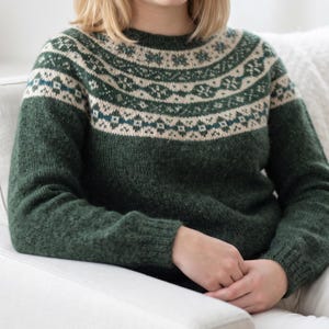 Pode incluir: Uma camisola de malha de manga comprida num tom verde floresta profundo. A peça apresenta um decote redondo e um jugo decorado com uma faixa horizontal de padrões geométricos intrincados em creme e azul-petróleo sobre uma base verde.