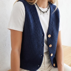 May include: A dark blue knitted waistcoat featuring a V-neckline and a front button closure with four light-coloured wooden buttons. The waistcoat has a vertical ribbed texture and is worn over a plain white short-sleeved t-shirt with a silver-toned necklace.
