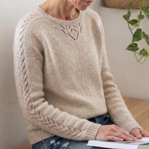 May include: A beige long-sleeve knit sweater featuring a round neckline with a delicate lace-like cutout detail and intricate cable knit patterns running down the length of each sleeve, finished with ribbed cuffs and a ribbed hemline.