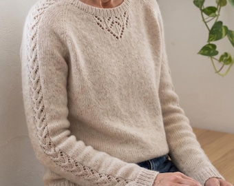 Puede incluir: Un suéter de punto beige de manga larga con cuello redondo, un delicado detalle calado en el escote y complejos patrones de trenzas a lo largo de las mangas, terminado con puños y dobladillo acanalados.