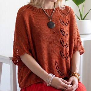 May include: A rust-colored knit top featuring an intricate leaf-patterned lace design, a wide boat neckline, and short, loose sleeves. The garment is styled with a dark circular pendant on a beaded chain and two clear, textured bracelets.