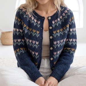 Op de afbeelding: Een marineblauw gebreid vest met een herhalend patroon van lichtroze en mosterdgele tulpen met saliegroene stelen. Het kledingstuk heeft een ronde halslijn, lange mouwen met geribbelde boorden en een knoopsluiting aan de voorzijde.