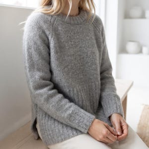 May include: A long-sleeved, heather gray knit sweater featuring a ribbed crew neckline, ribbed cuffs, and a ribbed hem. The sweater has a relaxed fit with raglan sleeves and side slits at the bottom hem, crafted from a textured, soft-looking yarn.