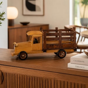 May include: A wooden toy truck featuring a light-toned cab and a dark-toned slatted cargo bed. The vehicle has four black wheels, a steering wheel visible inside the cab, and two small circular headlights on the front bumper.