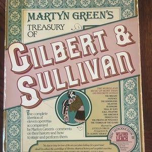 May include: A hardback book titled Martyn Green's Treasury of Gilbert & Sullivan with a cream, green, and maroon cover. The front features decorative borders, a central illustration of two figures in traditional attire, and text listing operettas.
