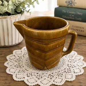 May include: A mustard-brown ceramic creamer pitcher featuring a glossy finish and a distinct chevron-patterned embossed design on the exterior. The pitcher has a wide pouring spout and a sturdy side handle, showcasing a uniform color throughout.