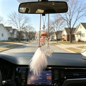 May include: A car rearview mirror hanging ornament featuring a small glass bottle with a wooden cap containing pink flowers, a silver wire crescent moon, a silver wire star, and a white feather, all suspended from thin brown cords.