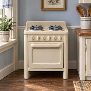 May include: A miniature cream-colored toy stove featuring four dark gray burners on the stovetop, six small control knobs along the front panel, and a central oven door with a horizontal handle, designed for a dollhouse kitchen setting.