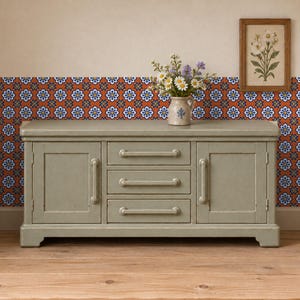May include: A sage green wooden sideboard featuring two side cabinets and three central drawers with matching horizontal handles. A ceramic pitcher filled with white and purple flowers sits on top, and a framed botanical print hangs above.