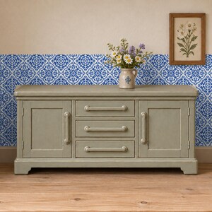 May include: A light gray wooden sideboard featuring two side cabinets and three central drawers with matching horizontal handles. A ceramic pitcher filled with white and purple flowers sits on top, next to a framed floral print.