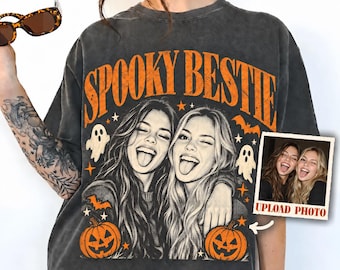 May include: A charcoal gray t-shirt featuring the orange text SPOOKY BESTIE in an arched font. The graphic includes a black and white illustration of two people with tongues out, surrounded by orange ghosts, bats, stars, and jack-o'-lanterns.