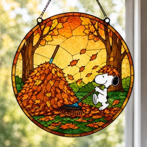May include: A circular stained glass suncatcher featuring Snoopy raking a large pile of orange and yellow autumn leaves. The scene includes brown tree trunks, green grass, and falling leaves, all framed by a dark border with a hanging chain.