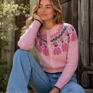 Peut inclure: Un cardigan en tricot rose clair avec une encolure ronde, des manches longues et une fermeture boutonnée sur le devant. Le vêtement est orné d'un motif floral bleu foncé et magenta sur la poitrine et les épaules, avec des bords-côtes.