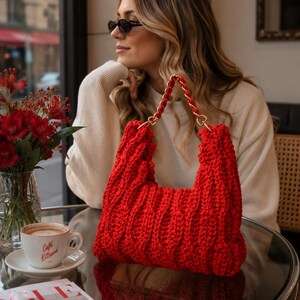 Peut inclure: Un sac à main rouge vif crocheté à la main, doté d'un motif en tricot torsadé épais et texturé, ainsi qu'une bandoulière en chaîne métallique dorée entrelacée de fil rouge assorti. Le sac présente une forme rectangulaire structurée.