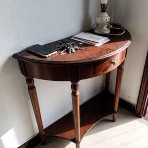 May include: A semi-circular wooden console table with a single drawer featuring a brass knob, four slender legs, and a lower shelf. On the surface sits a smartphone, a metal keyring with multiple keys, a stack of white envelopes, and a table lamp.