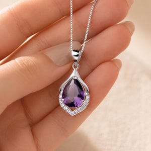 Puede incluir: Un collar de plata con un colgante de amatista morada en forma de pera. El colgante está enmarcado por un engaste de plata puntiagudo con pequeñas piedras claras de corte redondo en el borde inferior, suspendido de una fina cadena de plata.