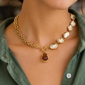 May include: A gold-toned necklace featuring a thick chain on one side and irregular white baroque pearls on the other. The necklace includes a central gold toggle clasp with a dangling, dark amber-colored teardrop pendant encased in gold.