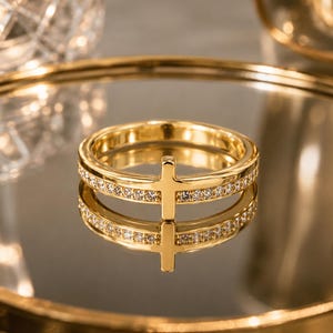 Könnte beinhalten: Ein goldfarbener Ring mit einem zentralen Kreuzmotiv und einem Band, das mit kleinen, klaren, funkelnden Steinen besetzt ist. Der Ring ist hochglänzend poliert und reflektiert das Licht auf seiner glatten Oberfläche und den facettierten Steinen.