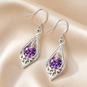 May include: A pair of silver-toned drop earrings featuring a teardrop-shaped frame with intricate filigree detailing at the base. Each earring holds a round, faceted purple gemstone suspended in the center, attached to a simple silver hook wire.