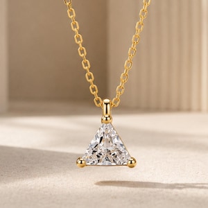 May include: A gold chain necklace featuring a triangular pendant with a clear, faceted stone. The pendant is held by three small gold prongs at each corner, suspended from a delicate gold link chain that catches the light with a polished finish.