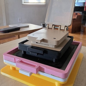 Op de afbeelding: Een meerlaags, stapelbaar persgereedschap met een felgele basis, een witte tussenlaag, een roze frame, een zwart rechthoekig onderdeel en een beige bovengedeelte met een scharnierend deksel dat naar boven toe openklapt.