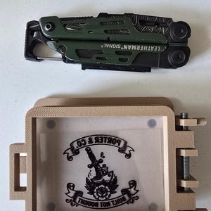 Może przedstawiać: Wielofunkcyjne narzędzie Leatherman Signal w kolorze oliwkowozielonym i czarnym, umieszczone nad beżową, wydrukowaną w 3D prostokątną ramką z przezroczystą wkładką. Wkładka zawiera czarną grafikę sztyletu w róży z napisem "BOKER & CO" oraz "BUILT NOT BOUGHT".