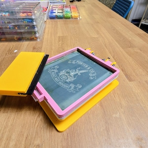 May include: A screen printing frame with a pink border and a yellow base, featuring a mesh screen with the white printed text Porter & Co. Built Not Bought. A yellow squeegee with a black rubber blade rests on the side of the frame.