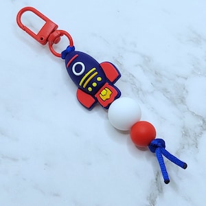Peut inclure: Un porte-clés en silicone en forme de fusée bleu et rouge avec une perle blanche et rouge et un cordon bleu.