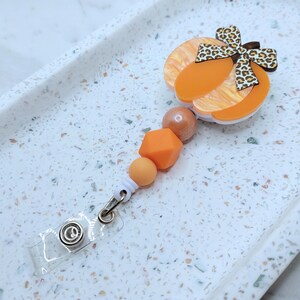 Op de afbeelding: Oranje en witte pompoenvormige hanger met een bruine en witte luipaardprint strik, bevestigd aan een wit en oranje kralenkoord met een transparante plastic badgehouder.