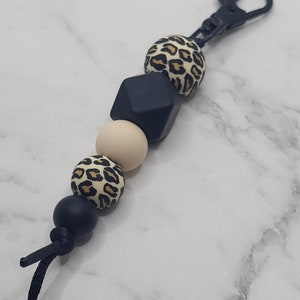 May include: A black and brown leopard print silicone keychain with black and beige beads. The keychain has a black metal clip.