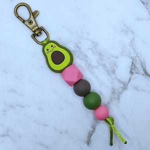 May include: A green avocado charm with a smiling face and a brown pit, attached to a gold key chain with pink, brown, and green beads.