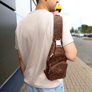May include: A brown leather sling bag with a distressed texture, featuring multiple zipped compartments, a wide shoulder strap, and a silver-toned metal buckle closure. The bag is worn across the chest, showcasing its compact design and functional hardware.
