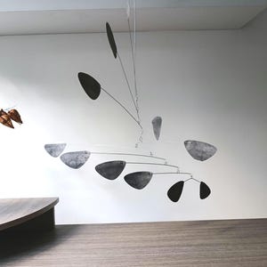 Puede incluir: Una escultura móvil moderna con un diseño minimalista. El móvil presenta varios elementos oscuros en forma de hoja suspendidos de finos alambres plateados. La escultura cuelga frente a una pared blanca y una superficie de madera.