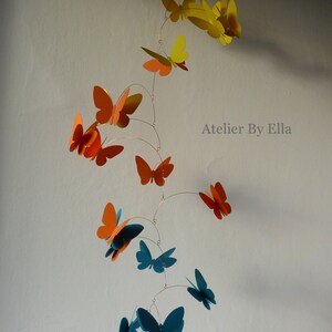 May include: A mobile with butterflies in shades of orange, yellow, and teal. The butterflies are suspended from a wire and move gently in the air. The mobile is labeled "Atelier By Ella".