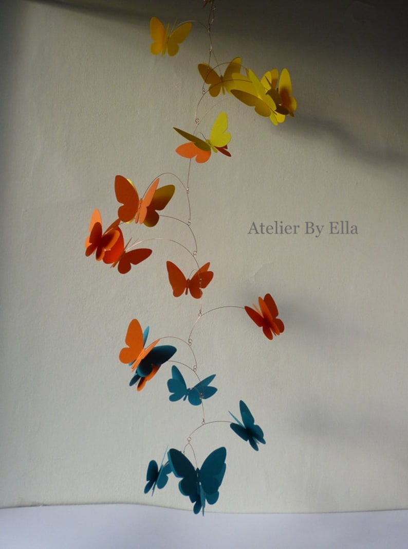May include: A mobile with butterflies in shades of orange, yellow, and teal. The butterflies are suspended from a wire and move gently in the air. The mobile is labeled "Atelier By Ella".