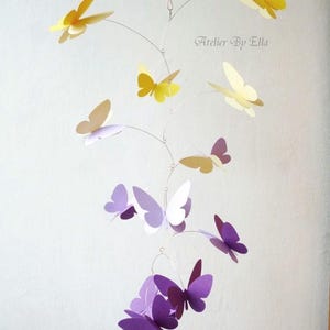 Puede incluir: Un móvil con mariposas de papel morado, blanco y amarillo colgando de un marco de alambre. El móvil está suspendido del techo. El texto "Atelier By Ella" es visible en la esquina superior derecha de la imagen.