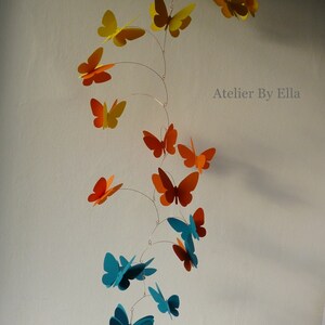 May include: A mobile with butterflies in shades of orange, yellow, and teal. The butterflies are suspended from a wire and hang at different lengths. The mobile is labeled "Atelier By Ella".