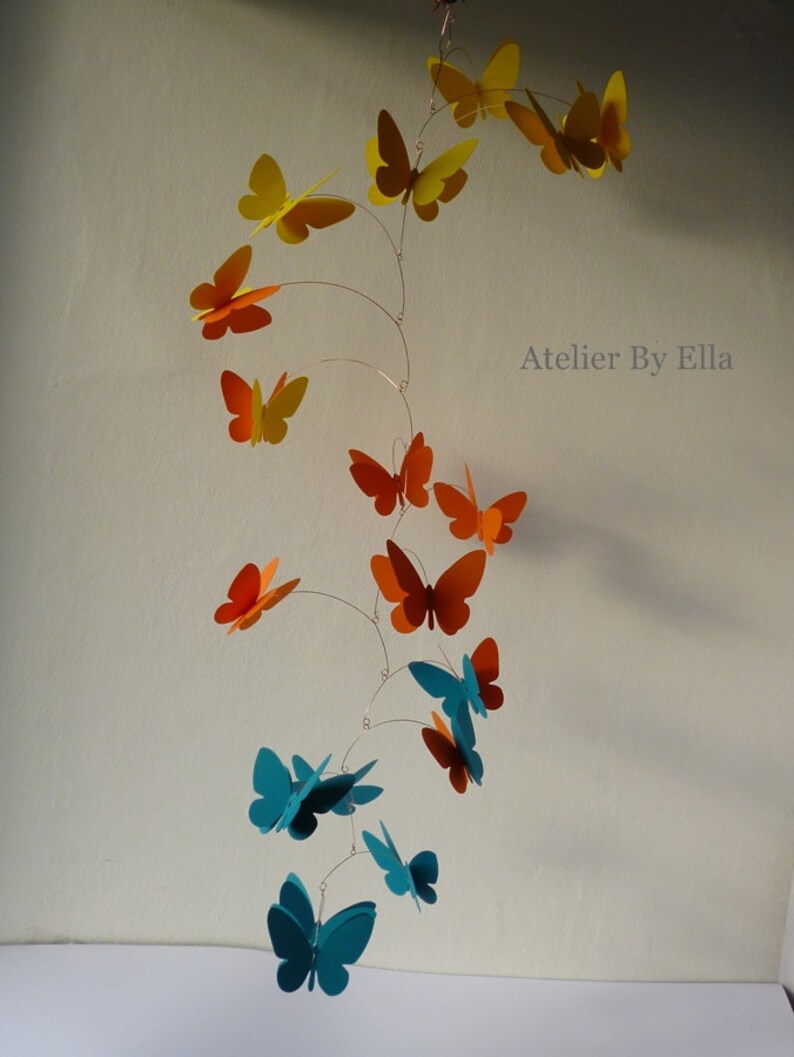 May include: A mobile with butterflies in shades of orange, yellow, and teal. The butterflies are suspended from a wire and hang at different lengths. The mobile is labeled "Atelier By Ella".