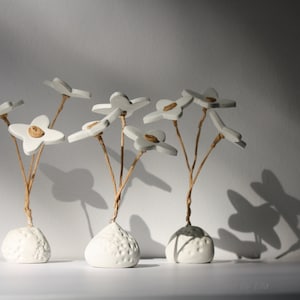 May include: Three white ceramic flower sculptures with brown stems. Each sculpture is in a white ceramic pot with a textured surface. The flowers are arranged in a cluster, with the stems extending upwards.