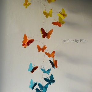 May include: A mobile with butterflies in shades of orange, teal, and yellow. The butterflies are suspended from a wire and hang at different lengths. The mobile is titled "Atelier By Ella".