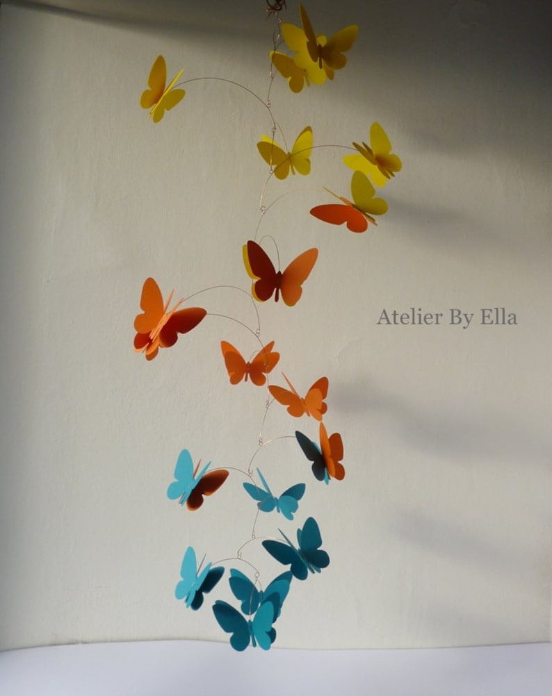 May include: A mobile with butterflies in shades of orange, teal, and yellow. The butterflies are suspended from a wire and hang at different lengths. The mobile is titled "Atelier By Ella".