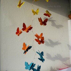 May include: A mobile with butterflies in shades of yellow, orange, and teal. The butterflies are suspended from a wire and hang at different levels. The mobile is hanging from the ceiling.