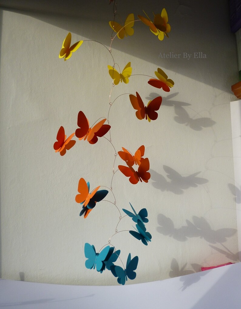 May include: A mobile with butterflies in shades of yellow, orange, and teal. The butterflies are suspended from a wire and hang at different levels. The mobile is hanging from the ceiling.