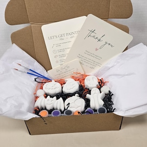May include: A cardboard box containing white plaster Halloween figures including pumpkins, a ghost, and a bat. The kit includes two blue-handled paintbrushes, small paint pots in orange, purple, and black, and printed cards with the text Let's Get Painting and Thank You.