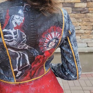 May include: A black and grey jacket featuring a hand-painted design on the back. The artwork depicts a skeleton figure on the left and a woman's face surrounded by a red and white sunburst pattern on the right. The jacket has yellow piping accents.