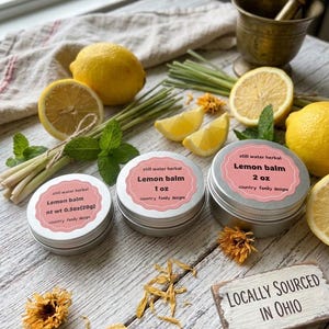 Könnte beinhalten: Drei runde silberne Metalldosen mit der Aufschrift still water herbal Lemon balm in den Größen 0,5 oz (20g), 1 oz und 2 oz. Die Dosen sind umgeben von frischen Zitronen, Zitronenscheiben, Zitronengras, Minzblättern und gelben Blütenblättern neben einem Schild mit der Aufschrift LOCALLY SOURCED IN OHIO.