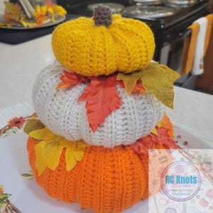 May include: A stack of three crocheted pumpkins in yellow, white, and orange, each topped with a small brown stem. The pumpkins are decorated with artificial autumn leaves in shades of red, orange, and yellow. A logo for RC Knots is visible.