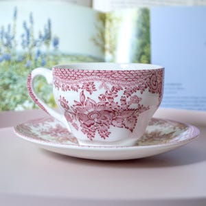 Peut inclure: Une tasse à thé et sa soucoupe assortie en céramique blanche, ornées d'un motif floral rouge foncé détaillé. Le design présente des illustrations botaniques complexes de fleurs et de feuilles, avec une bordure décorative en dentelle.