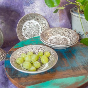 May include: A set of three round ceramic bowls featuring a brown and white transferware pattern of a landscape with trees and foliage. One bowl is filled with green grapes. The bowls have a decorative rim with a detailed floral and leaf motif.